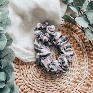 Könnte beinhalten: Ein lila-braunes, gemustertes Haargummi aus Stoff. Das Haargummi liegt auf einem geflochtenen Stroh-Untersetzer.