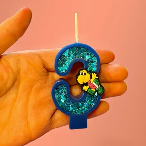 May include: Blue and green glitter birthday candle shaped like the number three with a small yellow turtle figurine on the side.