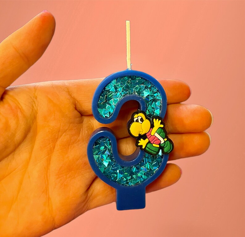 May include: Blue and green glitter birthday candle shaped like the number three with a small yellow turtle figurine on the side.