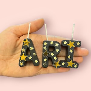 May include: Black candle letters spelling "ARI" with gold star accents. Each letter has a white wick.