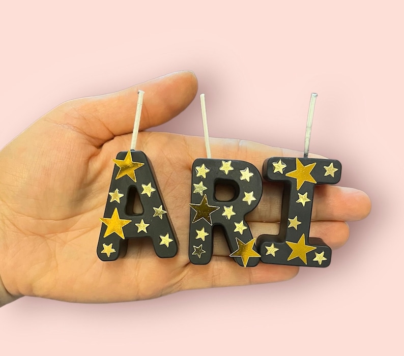 May include: Black candle letters spelling "ARI" with gold star accents. Each letter has a white wick.