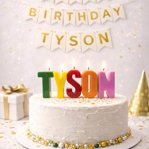 May include: A white frosted cake with colorful candles spelling "TYSON" and lit flames. A banner above reads "HAPPY BIRTHDAY TYSON". The cake is decorated with gold and white sprinkles. A gift box and party hat are in the background.