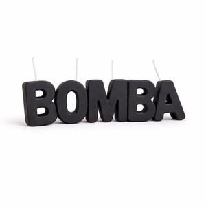 May include: Black candle set spelling out the word "BOMBA". Each letter is a separate candle with a white wick. The candles are thick and stand upright on a white surface.