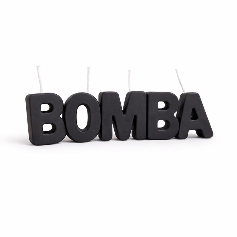 May include: Black candle set spelling out the word "BOMBA". Each letter is a separate candle with a white wick. The candles are thick and stand upright on a white surface.