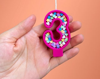 May include: Pink number three birthday candle with colorful sprinkles and stars.