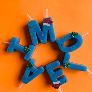 May include: Blue candle letters spelling "HOME" with a Santa hat and a Christmas tree on each letter. The candles are on an orange background.