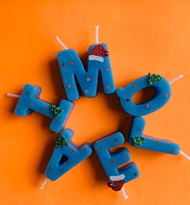 May include: Blue candle letters spelling "HOME" with a Santa hat and a Christmas tree on each letter. The candles are on an orange background.