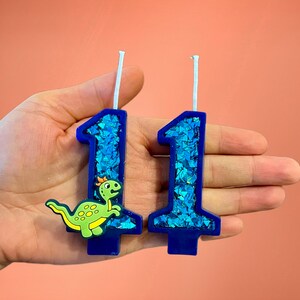 May include: Two blue birthday candles with glitter, one shaped like the number 1 and the other shaped like the number 1 with a green dinosaur on it.