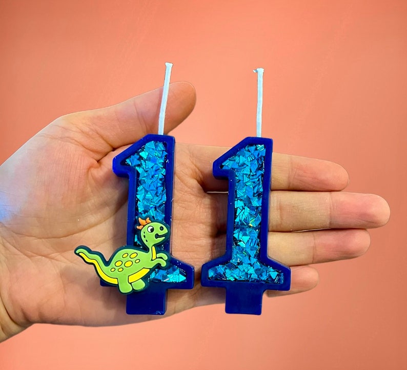 May include: Two blue birthday candles with glitter, one shaped like the number 1 and the other shaped like the number 1 with a green dinosaur on it.