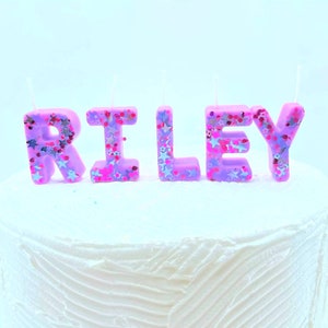 May include: Purple letter candles spelling out the name "RILEY" with pink and silver glitter. The candles are on a white frosted cake.