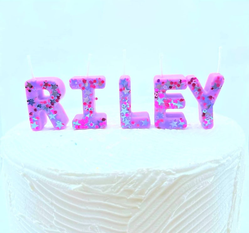May include: Purple letter candles spelling out the name "RILEY" with pink and silver glitter. The candles are on a white frosted cake.