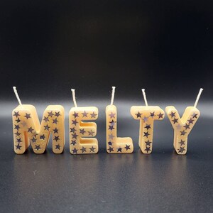May include: Five gold-colored candles spelling out the word "MELTY" with silver glitter stars. The candles are on a black background.