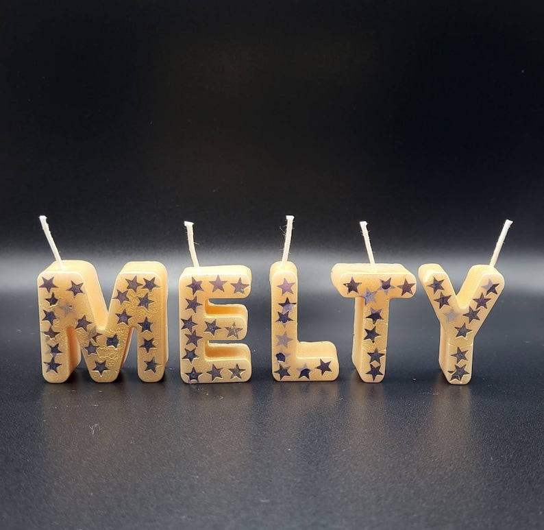 May include: Five gold-colored candles spelling out the word "MELTY" with silver glitter stars. The candles are on a black background.