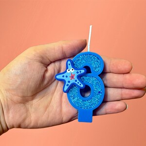 May include: Blue glitter birthday candle shaped like the number six with a blue and red starfish on the side.