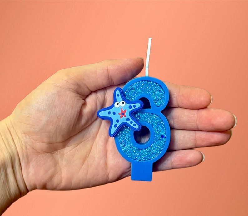 May include: Blue glitter birthday candle shaped like the number six with a blue and red starfish on the side.