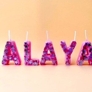 May include: Pink birthday candles spelling out the name "ALAYA" with silver glitter accents.