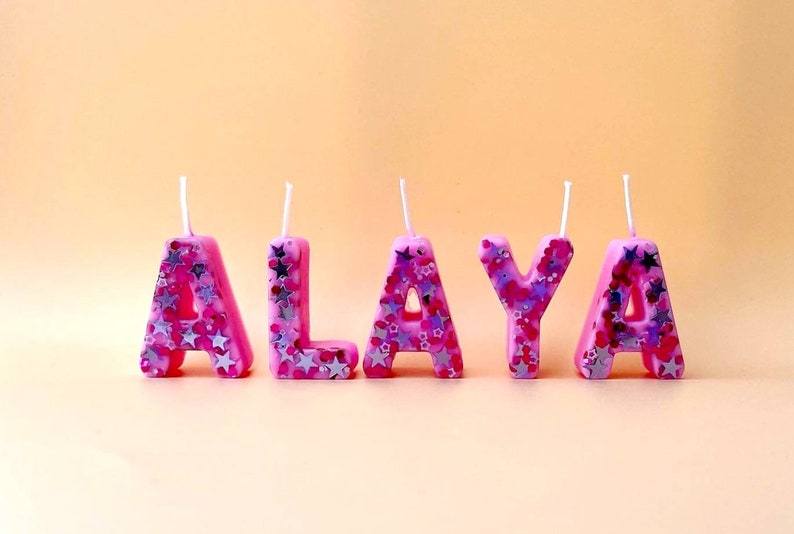 May include: Pink birthday candles spelling out the name "ALAYA" with silver glitter accents.