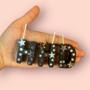 May include: Black letter candles with silver star accents spelling out "Happy".