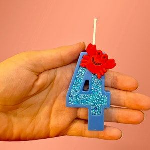 May include: Blue number four birthday candle with a red crab on top. The candle is covered in blue glitter.