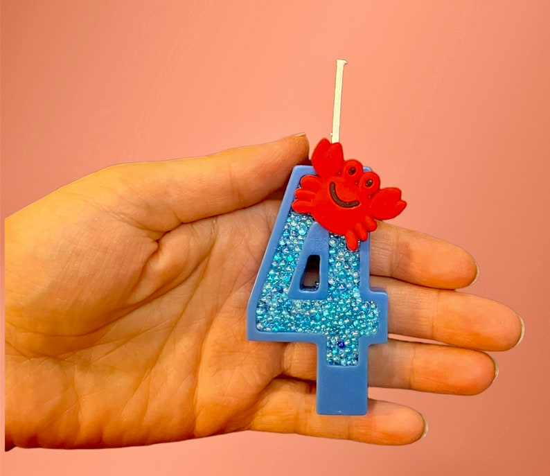 May include: Blue number four birthday candle with a red crab on top. The candle is covered in blue glitter.