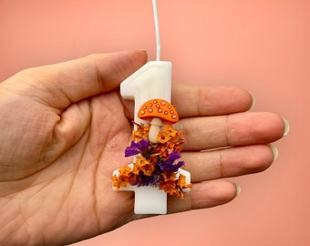May include: White candle shaped like the number one with a small orange mushroom and purple and orange flowers on top.