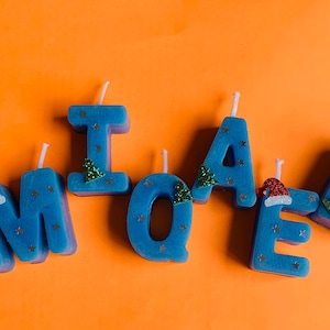 May include: Blue candle letters spelling out the name "MICHAEL" with gold glitter stars and green Christmas tree decorations. Some letters have red Santa hats.
