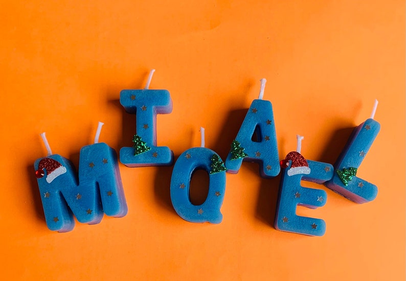 May include: Blue candle letters spelling out the name "MICHAEL" with gold glitter stars and green Christmas tree decorations. Some letters have red Santa hats.