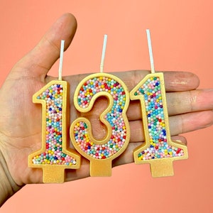 May include: Three gold-colored birthday candles with colorful sprinkles, shaped like the numbers 1, 3, and 1.