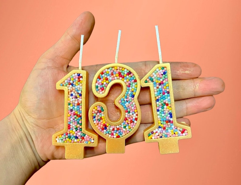 May include: Three gold-colored birthday candles with colorful sprinkles, shaped like the numbers 1, 3, and 1.