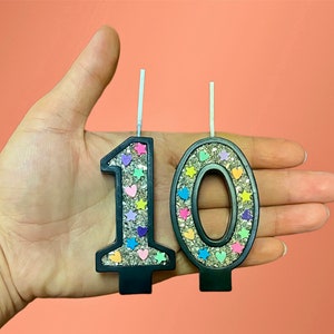 May include: Two black glitter birthday candles, one shaped like the number 1 and the other shaped like the number 0. The candles are decorated with colorful hearts and stars.