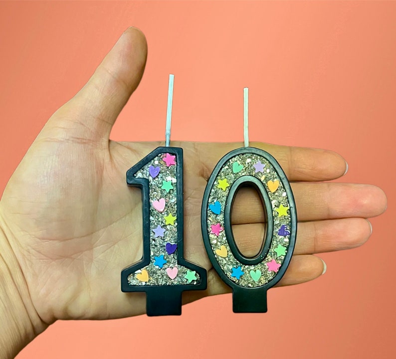 May include: Two black glitter birthday candles, one shaped like the number 1 and the other shaped like the number 0. The candles are decorated with colorful hearts and stars.