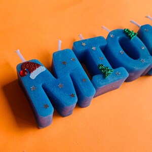 May include: Blue candle letters spelling out the name "MIKE" with silver glitter stars and green glitter Christmas trees. The letter "M" has a red Santa hat on top.