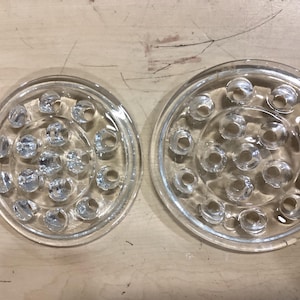 May include: Two clear glass flower frogs with multiple holes for arranging stems. The circular frogs are designed to hold flowers in place. The glass is transparent, allowing the stems to be visible.