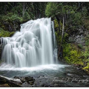 May include: A scenic photograph of a waterfall cascading over rocks into a pool of water. The waterfall is surrounded by lush green trees and moss-covered rocks. The image is a long exposure, creating a soft, blurred effect on the water.