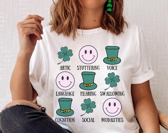 May include: White t-shirt with a graphic of green shamrocks and smiling faces with the words "Artic, Stuttering, Voice, Language, Hearing, Swallowing, Cognition, Social, Modalities"
