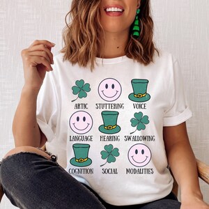 Puede incluir: Camiseta blanca con un gráfico de tréboles verdes y caras sonrientes con las palabras "Artic, Stuttering, Voice, Language, Hearing, Swallowing, Cognition, Social, Modalities"