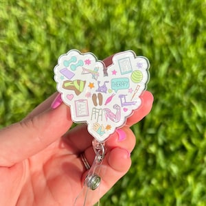 Puede incluir: Un portagafete blanco en forma de corazón con un diseño colorido de herramientas de fisioterapia y la palabra "THERAPY" en un bocadillo de diálogo. El portagafete está unido a un carrete de gafete retráctil de plástico transparente.