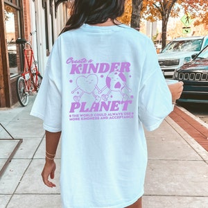 Op de afbeelding: Een lichtblauw T-shirt met een roze grafisch ontwerp met twee cartoonfiguren die hand in hand staan. De tekst op het shirt luidt "Create a Kinder Planet" en "The world could always use more kindness and acceptance."