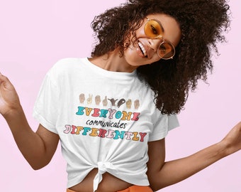 Puede incluir: Camiseta blanca con un gráfico de manos firmando en lenguaje de señas. El texto "EVERYONE communicates DIFFERENTLY" está impreso en letras de colores.