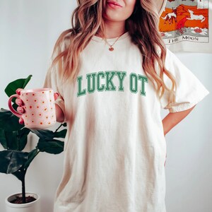 May include: A white t-shirt with the text "LUCKY OT" in green lettering. The shirt is oversized and has a relaxed fit.