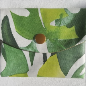 Op de afbeelding: Een kleine, witte stoffen tas met een groene en gele tropische bladerenprint. De tas heeft een gouden knoopsluiting.