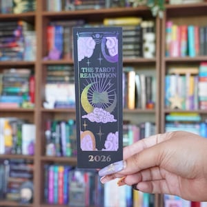 May include: A black bookmark with a celestial design featuring a crescent moon, clouds, and stars. The text "THE TAROT READATHON" and the year "2026" are printed on the bookmark. The background shows a bookshelf filled with books.