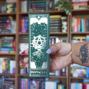 Puede incluir: Un marcapáginas verde con detalles plateados. Presenta un pentáculo, estrellas y la palabra «PENTACLES». El fondo es verde oscuro. El marcapáginas rectangular se sostiene en una mano, con una estantería de libros al fondo.