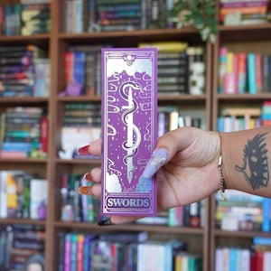 May include: A purple bookmark with a silver design featuring a sword, snake, and clouds. The word "SWORDS" is at the bottom. The background is a bookshelf filled with books.