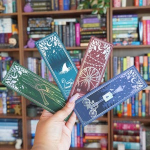 May include: Four rectangular bookmarks featuring intricate white line art designs. One is green with an axe, one is teal with a shark fin and float, one is maroon with a ferris wheel, and one is dark blue with a bicycle and street lamp.