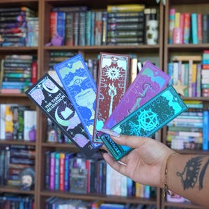 May include: A collection of five tarot-themed bookmarks with holographic accents. Each bookmark features unique designs, including celestial and mystical symbols, with the text "THE TAROT READATHON" on one. The bookmarks are held in front of a bookshelf filled with books.