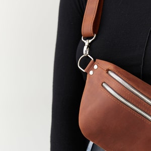 May include: A brown leather fanny pack with a silver buckle and zipper closures. The bag is worn across the body with a long brown leather strap.