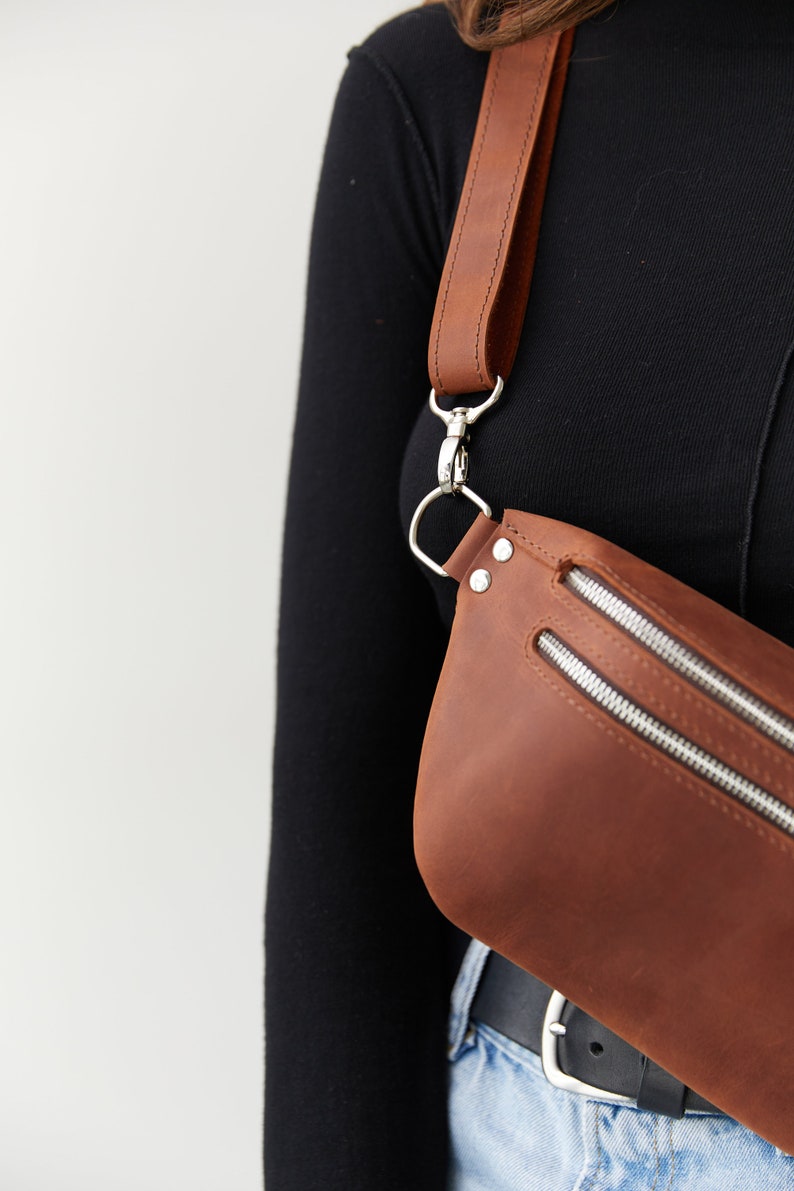 May include: A brown leather fanny pack with a silver buckle and zipper closures. The bag is worn across the body with a long brown leather strap.