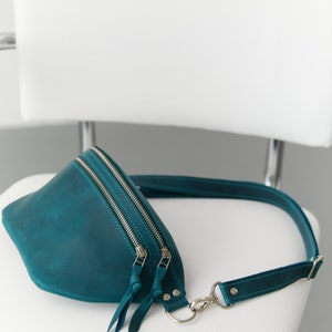 May include: Teal leather fanny pack with a silver zipper and adjustable strap. The bag is sitting on a white chair.