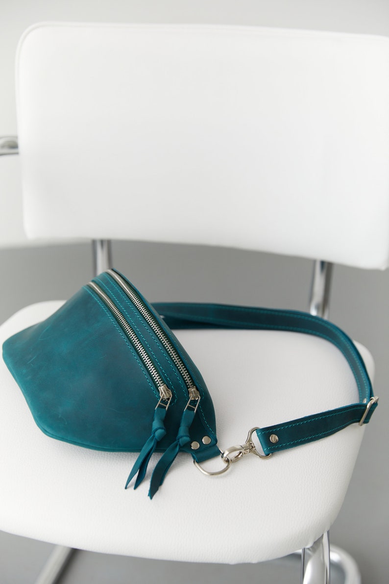 May include: Teal leather fanny pack with a silver zipper and adjustable strap. The bag is sitting on a white chair.